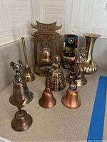Photo showing ten brass bells with various figures and designs along with a brass vase in background on countertop.