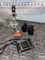 Photo of all items including Kodak Retina IIIc camera, HIT camera in case, microscope, and black binoculars arranged on carpet.