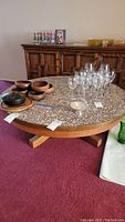 Photo showing full view of the round coffee table with cobblestone mosaic top, wooden bowls, and glassware on top, placed on red carpet with a dark wood credenza in the background.