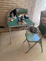 Photo showing metal folding table with green top and four matching folding chairs with green padding. Contents on table excluded from lot.