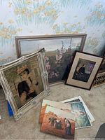 Three framed paintings displayed on the floor with five unframed prints stacked beside them. Framed art includes a figurative painting of a woman with a crown, a floral landscape, and a vintage-style portrait of a woman seated with a dark dress.