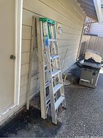 Two ladders leaning against an exterior wall next to a closed door and outdoor tools.
