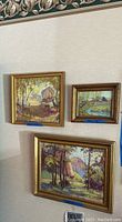 Three signed landscape paintings hung on a wall in inverted triangle arrangement