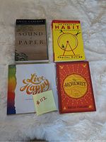 Four books laid out on a white furry background: Live Happy with a colorful cover, The Alchemist in red cover, The Power of Habit with a yellow cover, The Sound of Paper by Julia Cameron with a nature design cover.