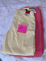 Two pairs of pants folded on a white surface, one yellow and one coral. Size tags visible, with yellow pants showing a sale tag.