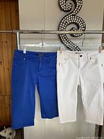 Two pairs of Tommy Bahama Bermuda jean shorts hanging side by side on hangers. Blue pair on left, white pair on right.