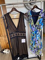 Two Max Studio dresses hanging on wooden hangers on metal bar. Left: black lace dress with nude lining. Right: floral flowing dress with mixed blues, purples, greens.