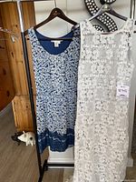 Two women's dresses hanging on a rack; left is a blue floral Max Studio dress with lace hem, right is a white lace sheer lined dress with tag indicating size L.
