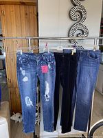 Front view of three women's jeans hanging on rack: distressed light blue with rips, black velvet feel, and classic blue denim