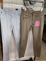 Photo of two pairs of women's designer jeans hanging side by side. Left is pale gray DL 1961 jeans, right is olive/khaki color Adriano Goldschmied jeans with multiple zippered pockets.