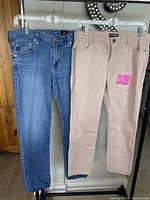 Front view of two pairs of jeans hanging, one blue denim Buffalo jeans and one powder pink Adriana Goldschmied jeans with size sticker.