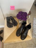 Pair of black patent leather thong sandals and pair of black leather Chelsea boots on a padded stool with purple flowers nearby.