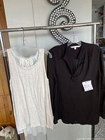 Two women’s designer tops hanging on clear hangers on a clothing rack, left is a sleeveless cream blouse with pleated collar, right is a black blouse with collar and tag attached.
