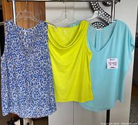 Three women's tops hanging on display, including a blue-white animal print sleeveless blouse, lime green tank, and light blue blouse.