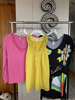 Three women's tops hanging on a silver rod: pink long sleeve, yellow sleeveless, and black floral sleeveless.