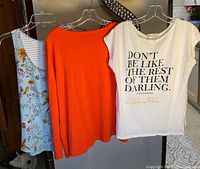 Three women's tops on hangers: blue floral tank, orange long sleeve top, white graphic tee
