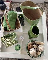 Top-down view of green tote, towels in basket, ceramic owl vase, black and green vase, glass bowl with decorative items, and small green glass votive holder arranged on white table.