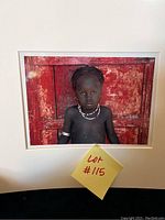 Color photo of the front of the photographer print showing a young Ethiopian child with jewelry against a red door background, mounted with white matting and lot number sticker