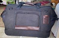 Front view of large black Samsonite duffle bag with leather accents and zippered front pocket, showing top handles and luggage hardware.