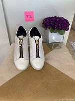 Front view of women's white leather sneakers with gold central zipper, paired and placed upright.