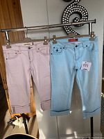 Front view of two pairs of women's crop jeans hanging on a rod inside a room, showing the folded cuffs and front pockets, one in subtle powder pink and the other in subtle turquoise.
