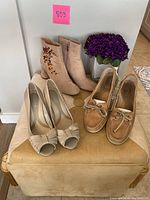 Three pairs of women's shoes including beige Sperry Topsider boat shoes, beige Zara zip ankle boots with floral embroidery, and cream Naturalizer heels displayed on beige stool with purple flowers.