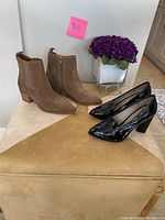 Both pairs of shoes side by side on a beige surface with purple flowers in the background, showing the taupe suede ankle boots and black patent leather heels.