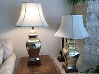 Two gold colored porcelain lamps on wooden side tables with white shades, one lamp turned on.