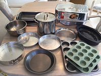 Full view of all the baking and cooking pans on a table showing the assortment of items including stock pot, roasting pan, cake pans, muffin tins, baking trays, mixing bowl, and ceramic dish.