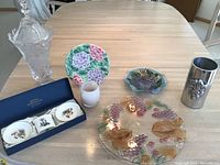 Overview of the lot on a table showing a large glass tray with grape design, small glass bowl with floral design, frosted glass candle holder, Royal Worcester bone china coasters in box, and silver-toned metal vase with grape motif.