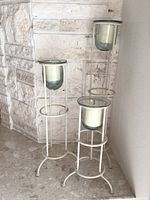 Three white metal candle holders with glass cups, different heights, shown standing near a stone wall.