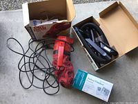 Handheld red Dirt Devil vacuum with power cord, original box in background, box of attachments visible