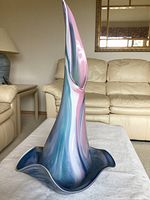 Tall glass art vase with twisted elongated shape, pink and blue swirls sitting on white surface with couch and lamp background.