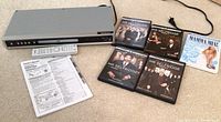 Full view of the Magnavox DVD player, remote control, owner's manual, and five DVDs displayed on carpet.