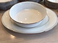 Close-up showing stacked white bowls, salad plates, and dinner plates with silver trim and scalloped edging representing the Noritake Sterling Cove pattern.