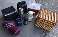 Photo showing woven picnic basket, black coolers, metal and plastic thermoses, wine bag, and drink holders grouped together on concrete.