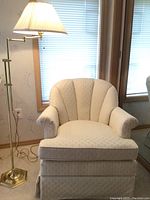 Front view of off white scalloped pattern upholstered rocking chair with rocking and spinning features next to the brass pivoting floor lamp with pleated white shade