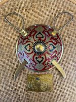 Front view of brass dhal shield with red and silver engraved pattern, two kirpans crossed and small brass plaque below