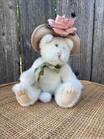 Front view of white Boyd's Bears Lola Ninelives plush bear sitting on a wicker surface, wearing beige hat with a pink flower and green ribbon tie at neck.