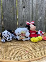 Two Disney plush toys side by side on a surface against a wooden fence background showing Dalmatian with Patch tag and Minnie Mouse with candy cane and red dress.