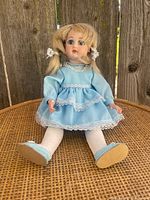 Photo of the vintage doll seated against a wooden backdrop showing the full figure in a blue dress with lace, white stockings, and blue shoes.
