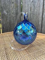 Blue glass hanging ornament with iridescent diamond pattern displayed on clear acrylic curved stand on wicker surface with wooden fence background.