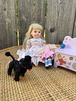 Photo shows Madame Alexander doll sitting with small rag doll, black toy poodle, and pink toy chest on wicker surface against wooden background.