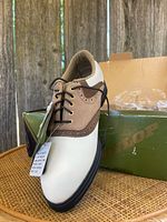 Single Dunlop golf shoe outside the box showing detail of white leather upper with brown leather accents, black soles, lace-up closure with gold eyelets, and stitching detailing.