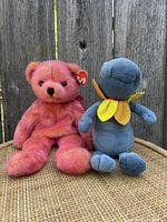 Front view of Rouge The Bear and Batty the Bat Ty Beanie Babies sitting on a woven rattan surface against a wooden fence background.