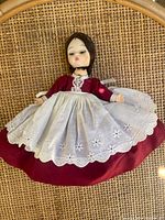 Full view of the vintage Alexander Kin doll laying on a woven surface showing the red dress and white embroidered apron.
