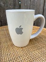 White ceramic coffee cup with gray Apple logo against a woven mat background, showing front with logo.