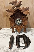 Front view of the vintage wooden Scottish cuckoo clock with carved bird and leaves and three pinecone-shaped weights and chains arranged in front.