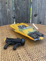 Yellow Star Wars Landspeeder toy with black blaster accessory shown on woven surface with wood fence background.