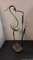 Full view of stone and metal bird sculpture showing shape and materials with stone body and metal neck, legs and cattails.